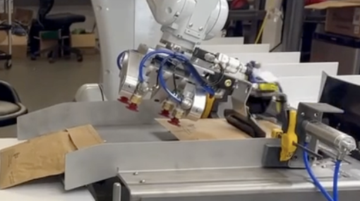 A robotic gripper placing a kraft mailer into a metal guide on a packing bench